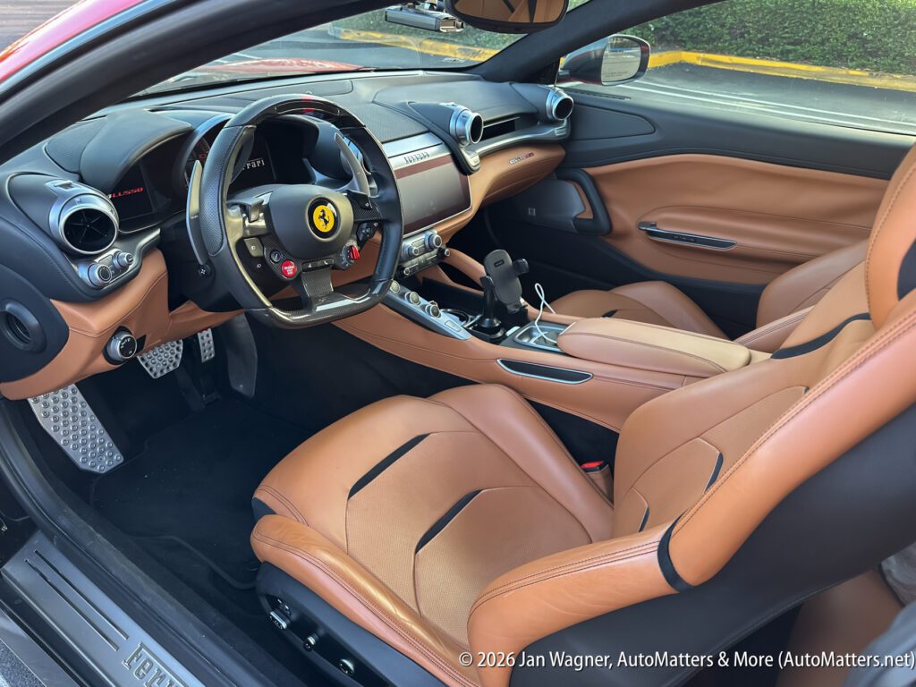 Interior of a Ferrari showing tan leather seats, a black steering wheel with controls, and a modern dashboard with metallic accents.
