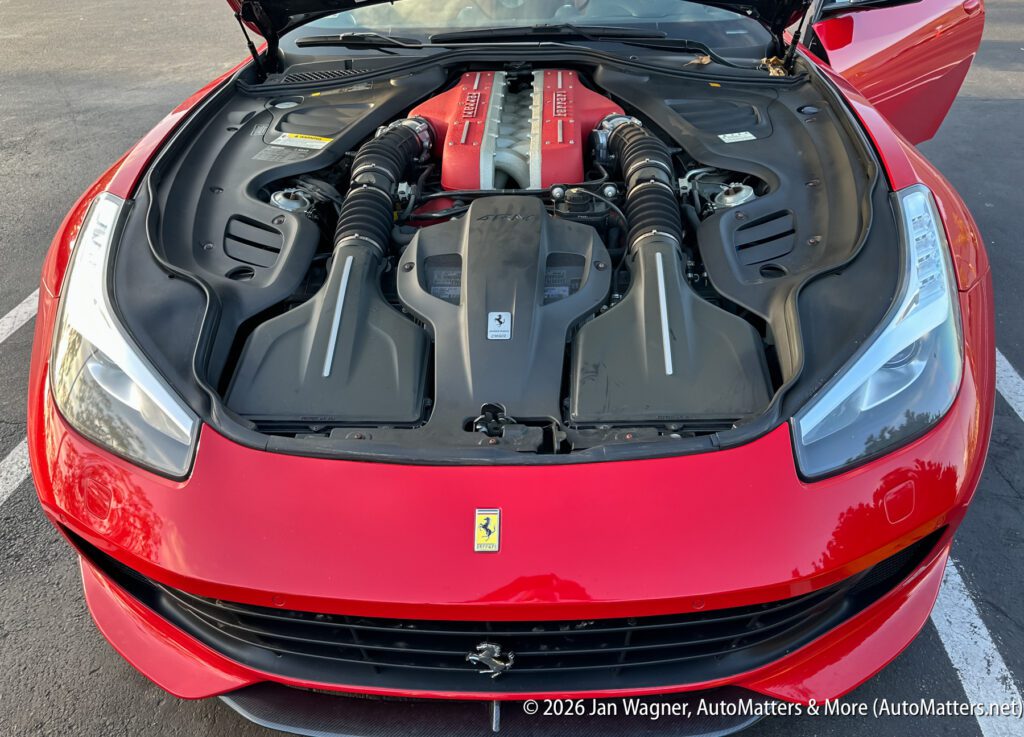 A red Ferrari with its hood open, revealing a clean V12 engine bay, parked on an asphalt surface.