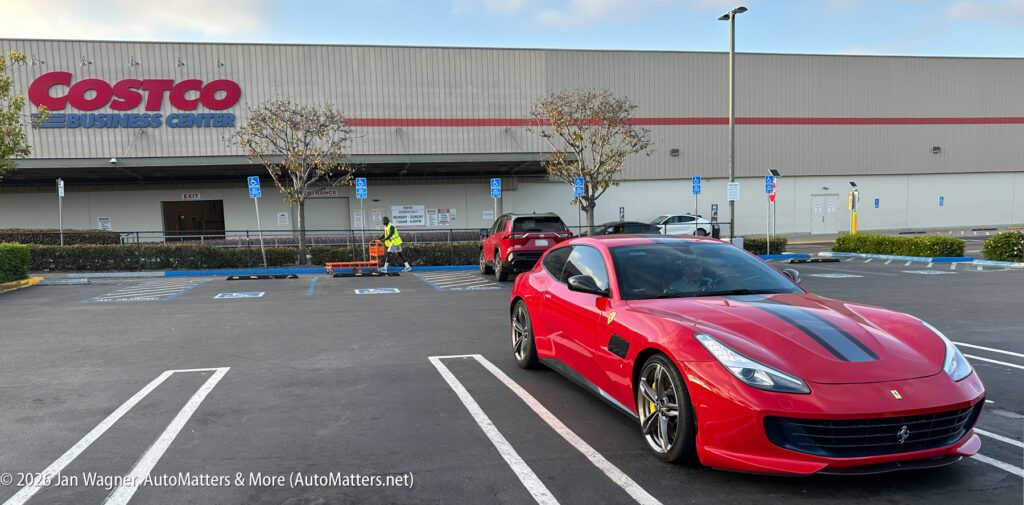 A red Ferrari is parked in a Costco Business Center parking lot with a red pickup truck and a worker pushing carts in the background.