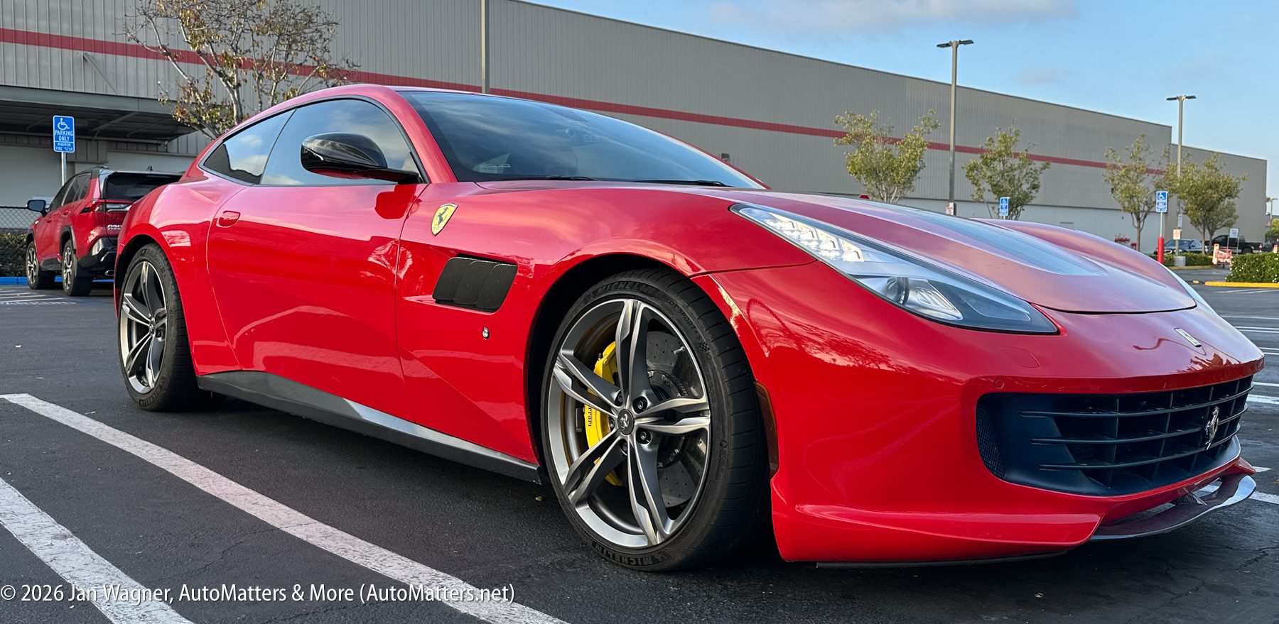 A red Ferrari sports car is parked in a lot near a warehouse-style building, with other vehicles and trees in the background.