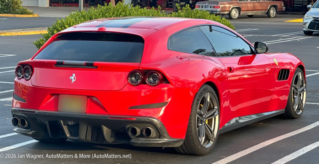 A red Ferrari GTC4Lusso parked in a parking lot, viewed from the rear left side, with a classic car in the background.