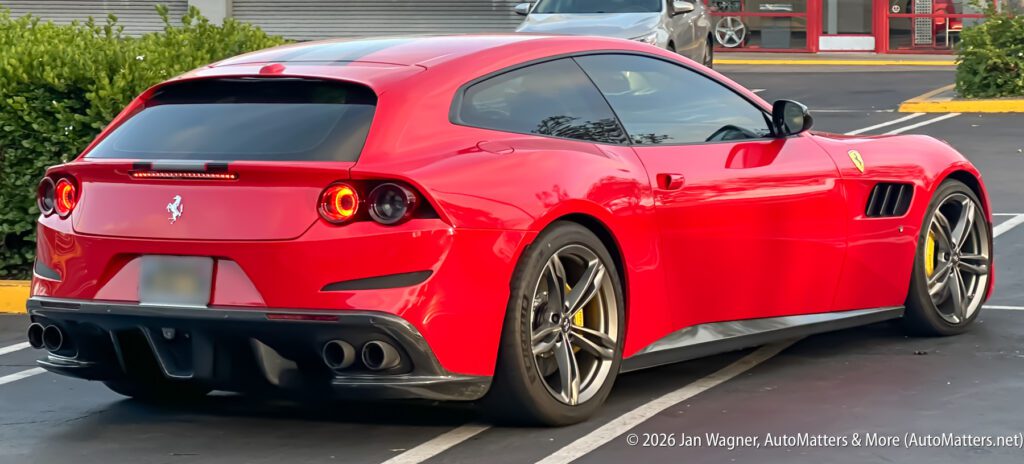 A red Ferrari sports car is parked in a lot, viewed from the rear left side. The car features yellow brake calipers and distinctive quad exhaust pipes.