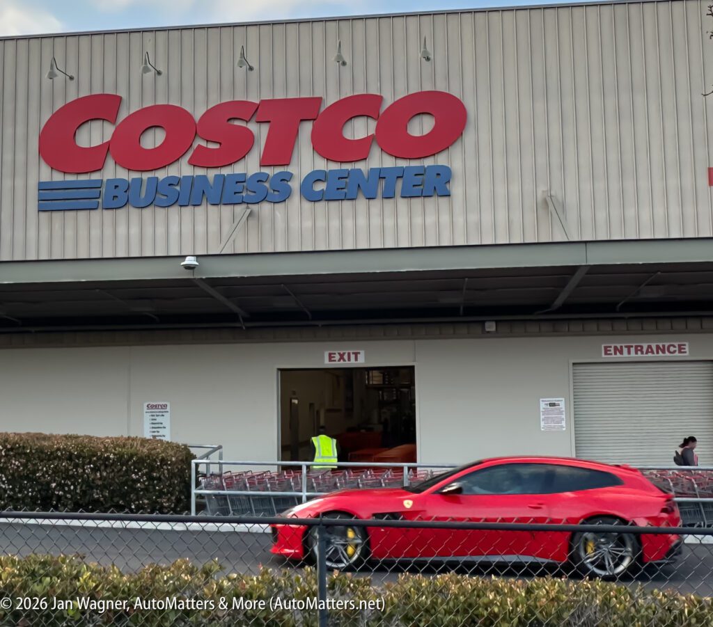 A red sports car passes in front of a Costco Business Center building, with shopping carts and an open exit door visible.