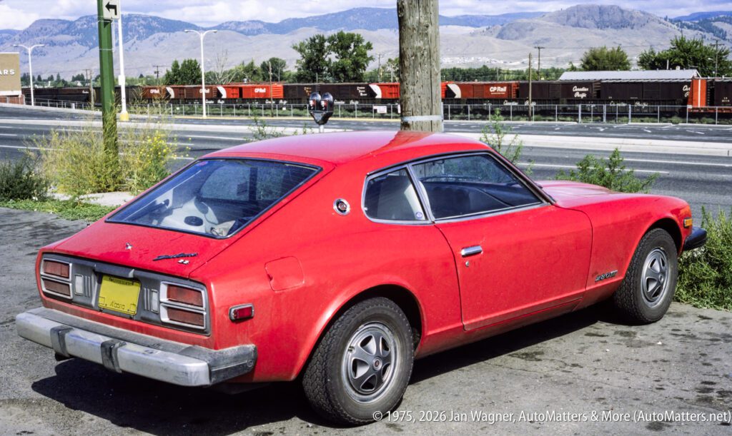 A red vintage Datsun 280Z is parked near a road with a freight train and mountains visible in the background.
