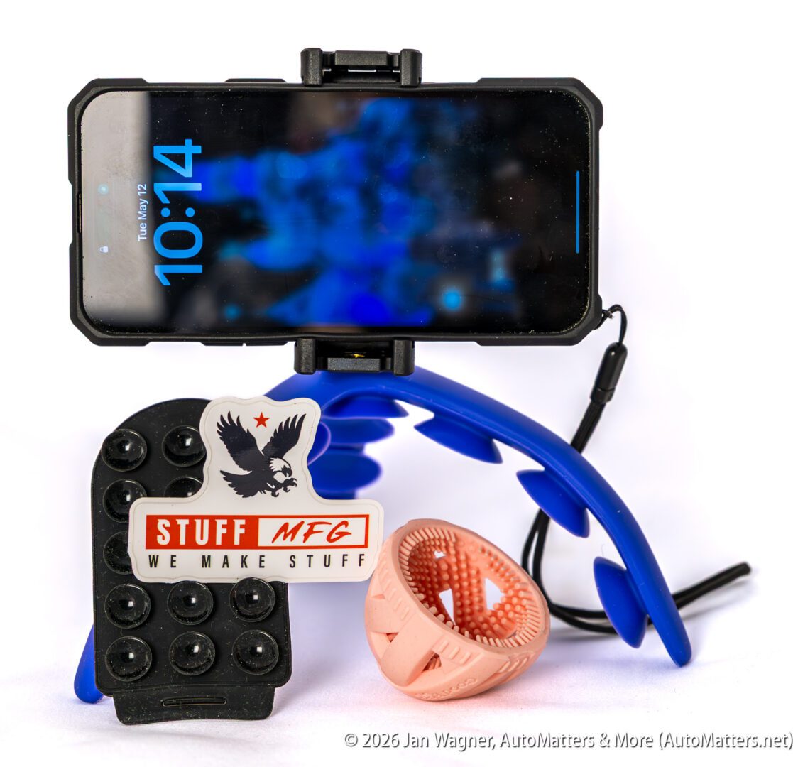 A smartphone in a black holder displays the time 10:14, surrounded by a blue flexible stand, a sticker reading "STUFF MFG," a suction cup mount, and an orange ring-shaped object.