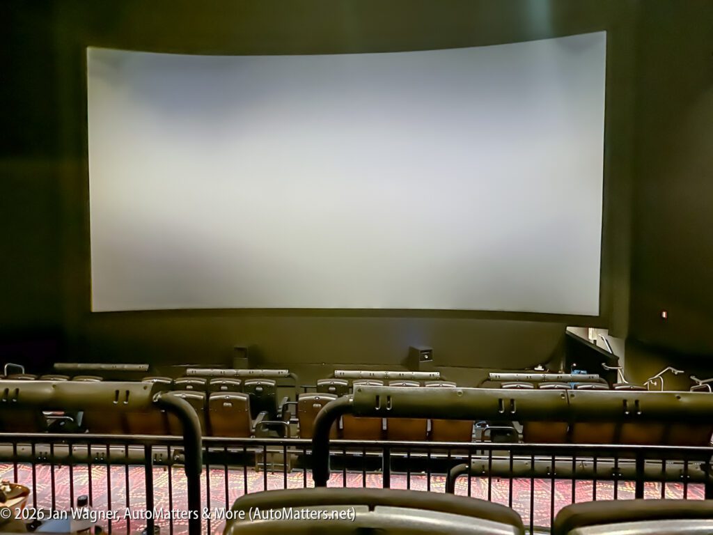 Empty movie theater with rows of seats facing a large blank screen; image taken from the back of the auditorium.