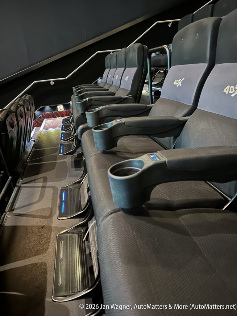 Row of black 4DX theater seats with cup holders and footrests, viewed from the side in a dimly lit cinema.