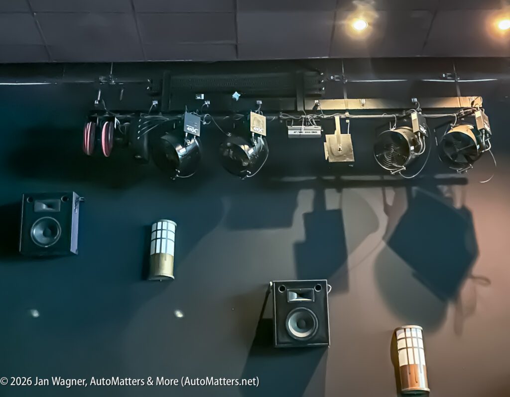 Ceiling-mounted stage lights, speakers, and wall sconces are installed on a dark wall, illuminated by overhead spotlights.