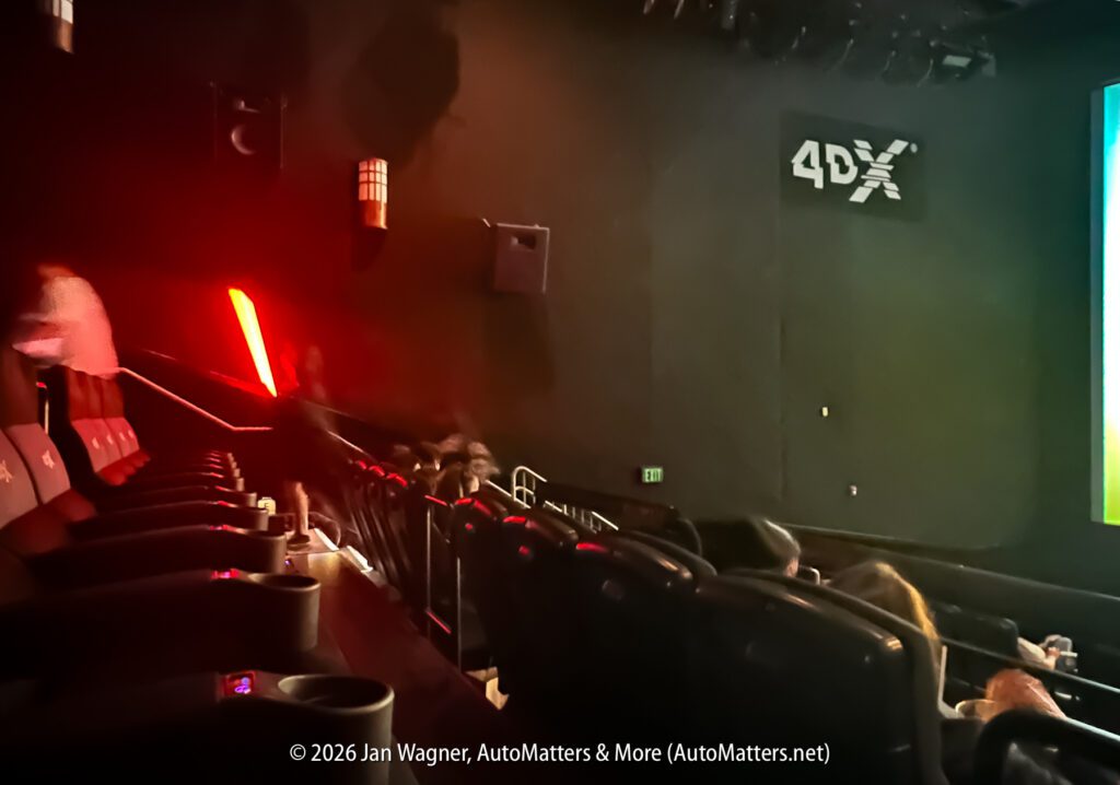 Interior of a 4DX movie theater with black seats and dim lighting; a person holding a red lightsaber replica is walking down the aisle.