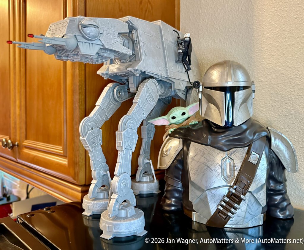A toy AT-AT walker stands next to a Mandalorian helmet bust with a small Grogu figure peeking from behind, displayed on a kitchen counter.