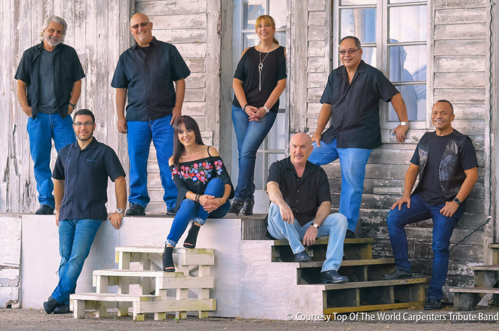 Seven people pose in front of a weathered wooden building, some sitting on steps and others standing, all dressed in casual attire.