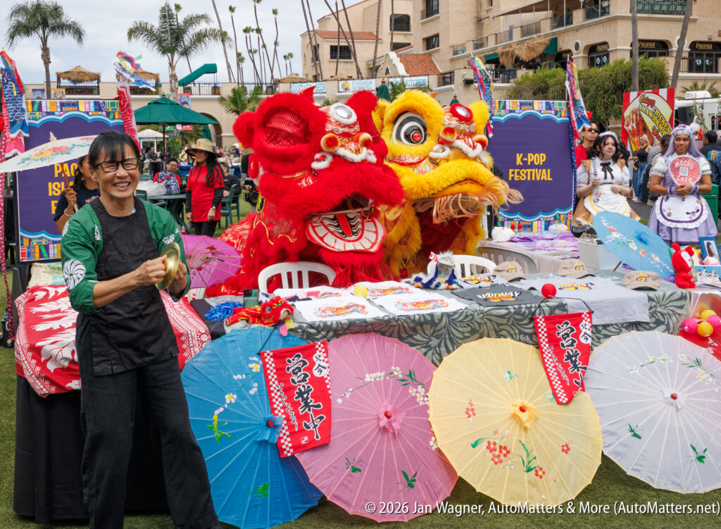 A woman stands smiling next to two lion dance costumes at a festival booth decorated with colorful umbrellas and banners, with people and tents in the background.