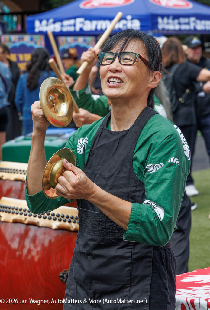 A woman in a green shirt and black apron plays small cymbals outdoors at a public event, smiling as she performs. Other people and tents are visible in the background.