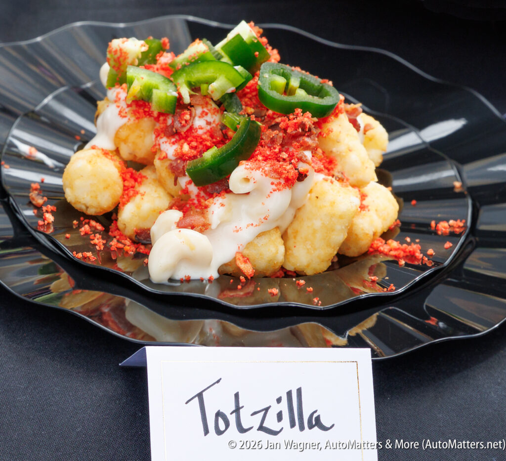 A black plate holds tater tots topped with white cheese sauce, chopped jalapeños, and red seasoning. A label in front reads "Totzilla.