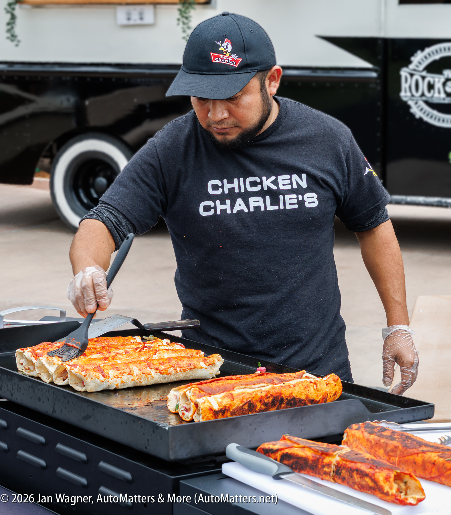 A man wearing a "Chicken Charlie's" shirt cooks large rolled tortillas on a flat top grill next to a food truck.