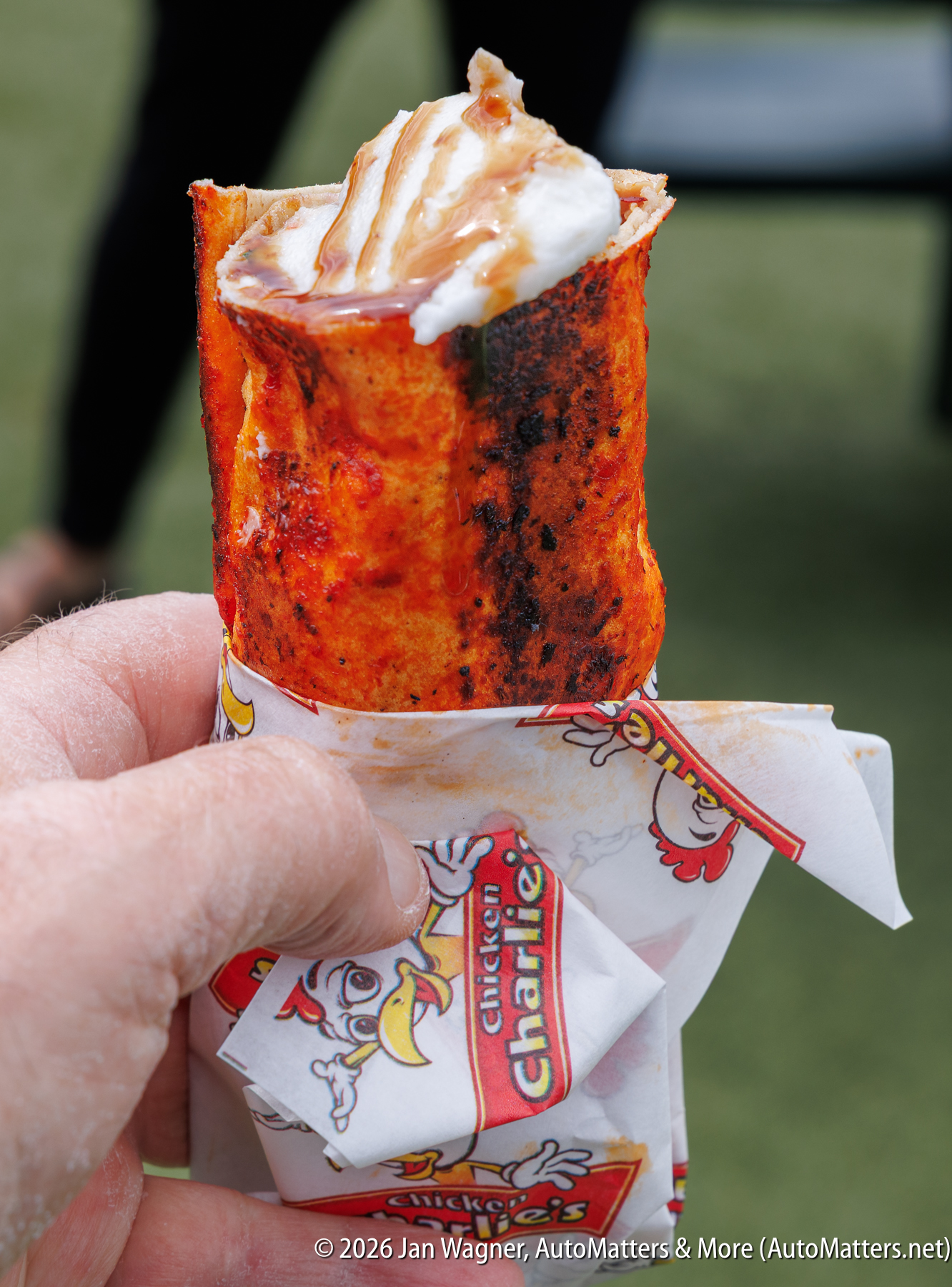 A hand holds a rolled, spicy orange tortilla cone filled with whipped cream, wrapped in Chicken Charlie's branded paper.