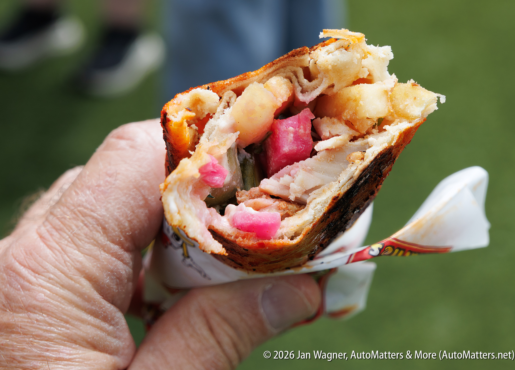 A hand holding a partially eaten wrap filled with sliced meat, pink pickled vegetables, potatoes, and other ingredients.