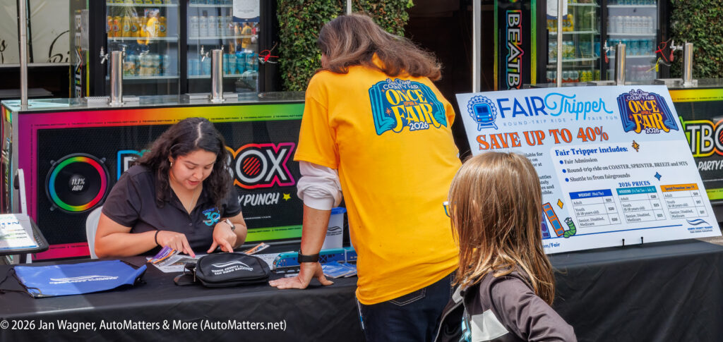 A woman sits at a booth handing out brochures as two people stand nearby; a display promotes fair tickets and discounts.