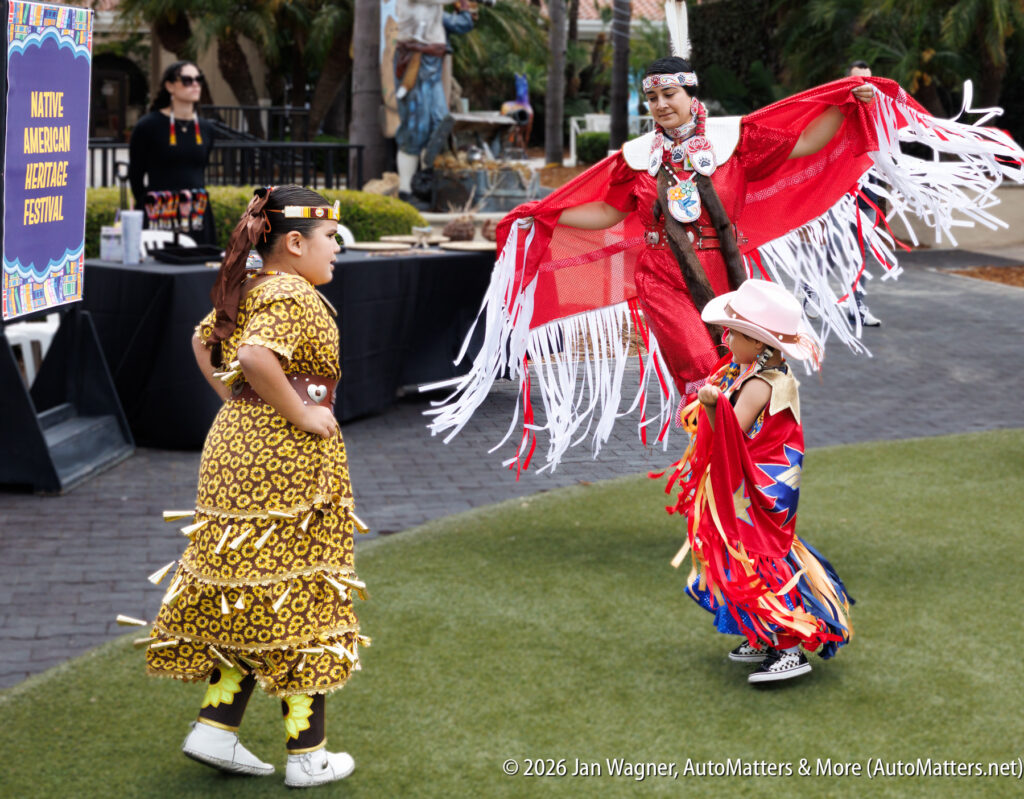 Three people in traditional Native American regalia perform a dance outdoors at a Native American Heritage Festival. A sign and greenery are visible in the background.
