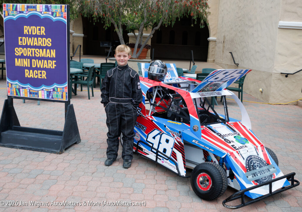 A young boy in a racing suit stands next to a small race car numbered 48, with a sign reading "Ryder Edwards Sportsman Mini Dwarf Racer" nearby.