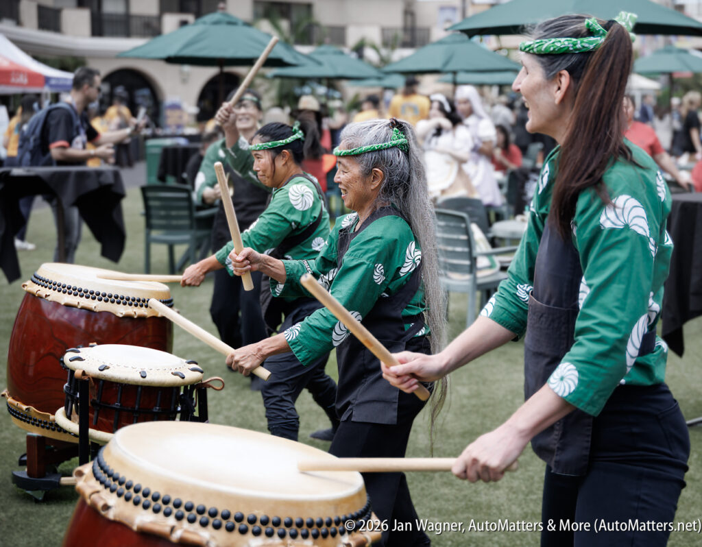 Three people in green attire play traditional Japanese drums outdoors, with an audience seated at tables in the background.