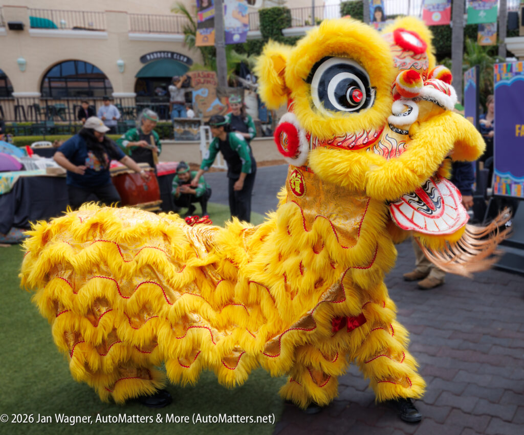A performer in a yellow lion dance costume participates in a cultural event outdoors, with people and festival decorations in the background.