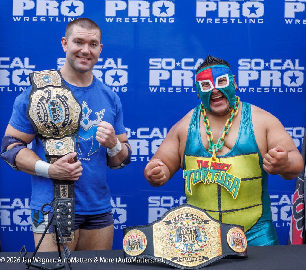 Two professional wrestlers stand side by side, smiling and holding championship belts, in front of a "SoCal Pro Wrestling" backdrop.