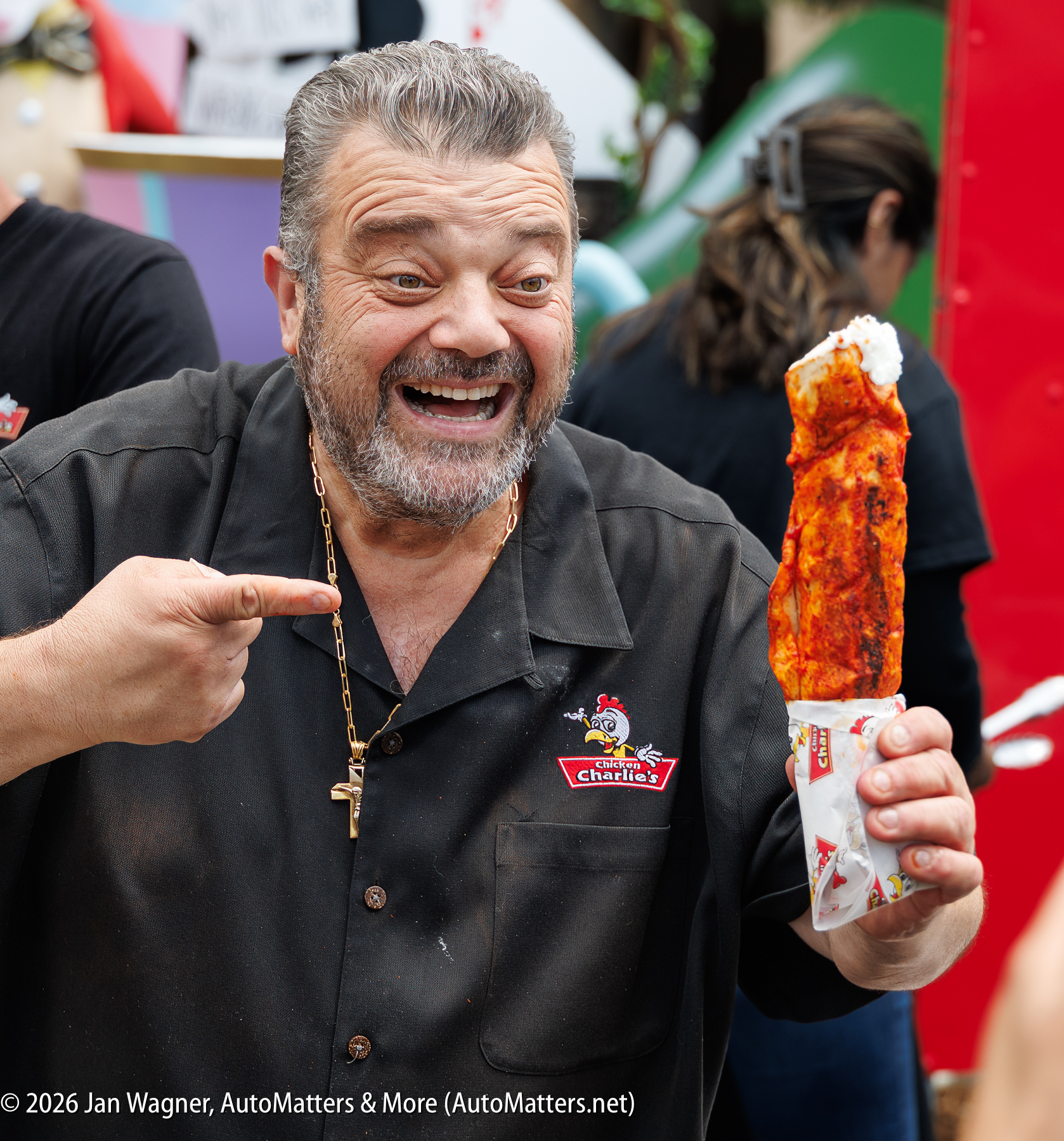A man in a black shirt smiles and holds a large, red, sauce-covered food item wrapped in paper.
