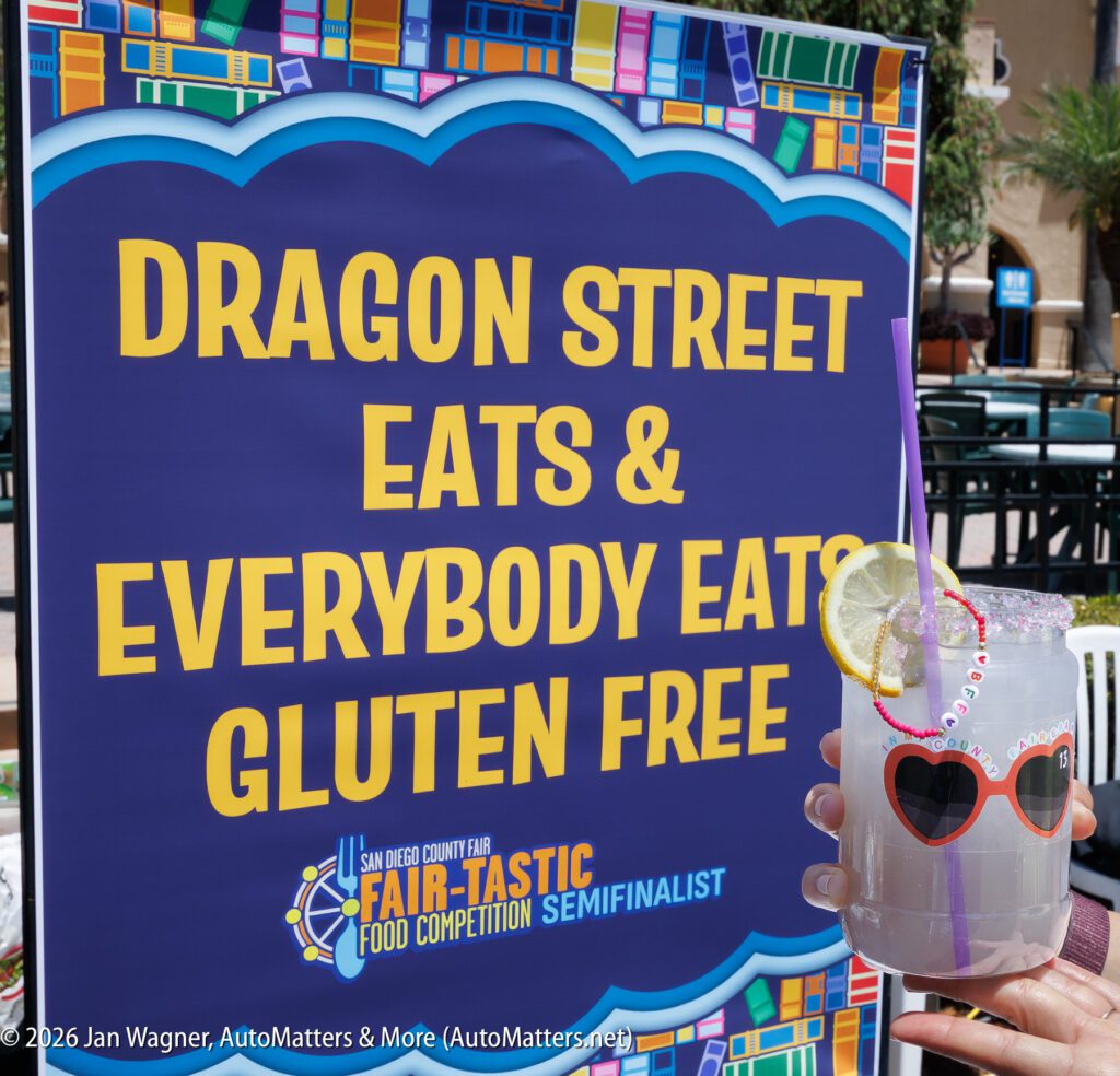 A sign reads "Dragon Street Eats & Everybody Eats Gluten Free" with a Fair-Tastic Food Competition logo; in front, a drink with a lemon slice, straw, and heart-shaped sunglasses is held.