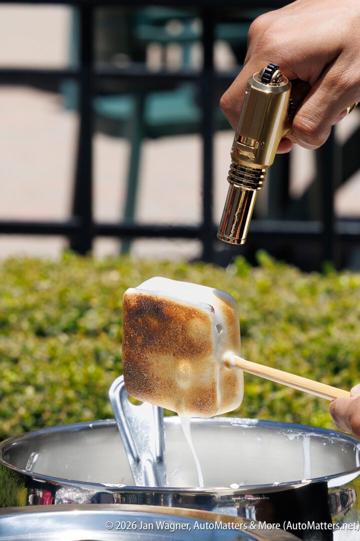A hand uses a small torch to toast a marshmallow on a skewer over a metal bowl, with melted marshmallow dripping down.