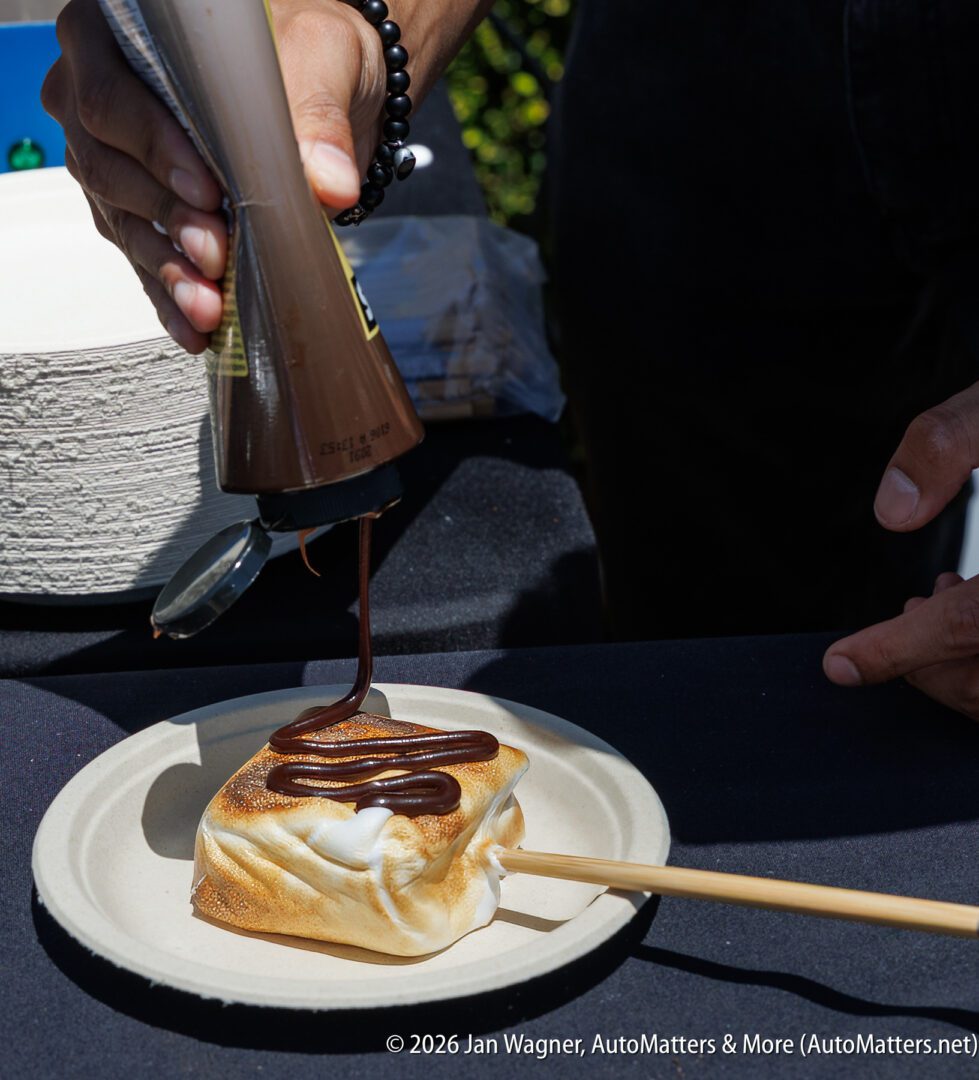 A person squeezes chocolate sauce onto a toasted marshmallow square on a stick, placed on a paper plate.