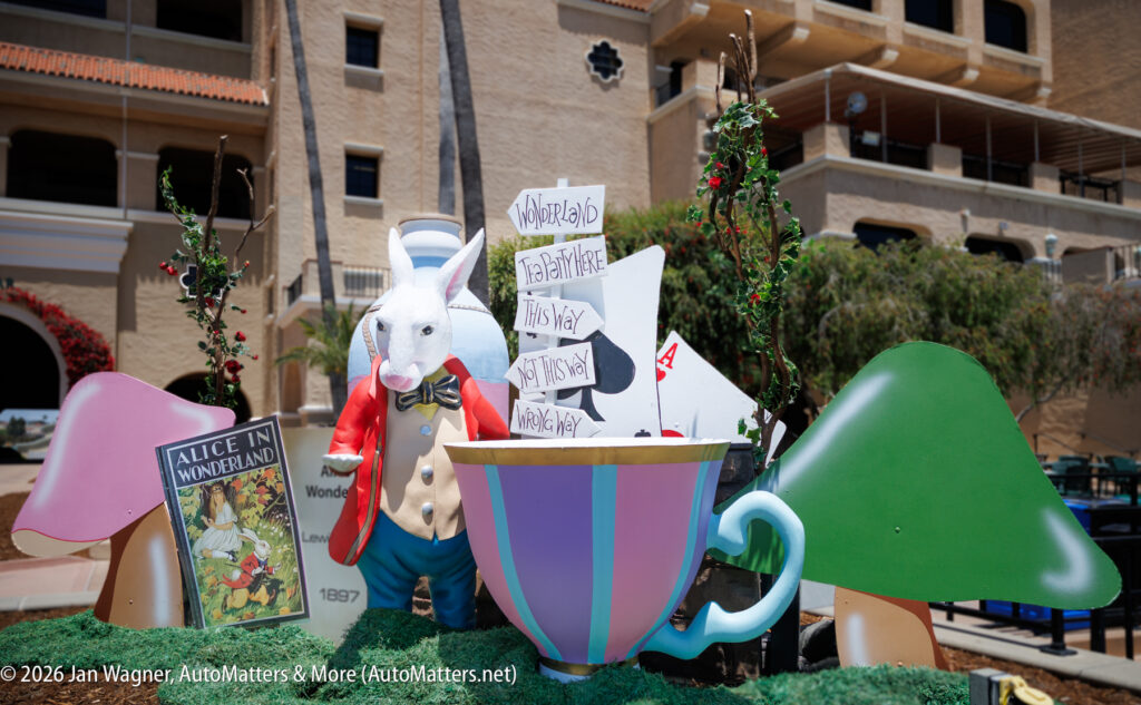 A decorative display with a large teacup, the White Rabbit, an "Alice in Wonderland" book, and a wooden signpost in front of a building.