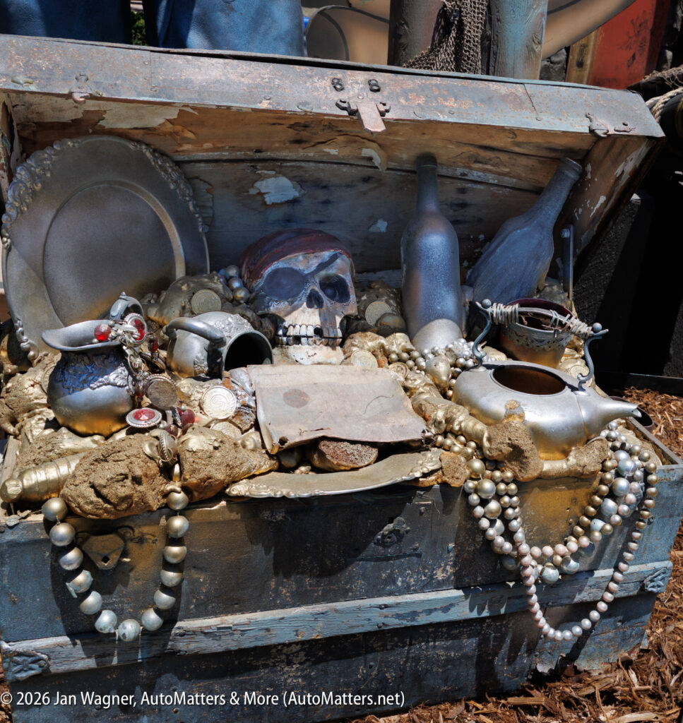 An open treasure chest filled with gold coins, pearls, jewelry, a metal skull with a red bandana, bottles, and various silver objects.