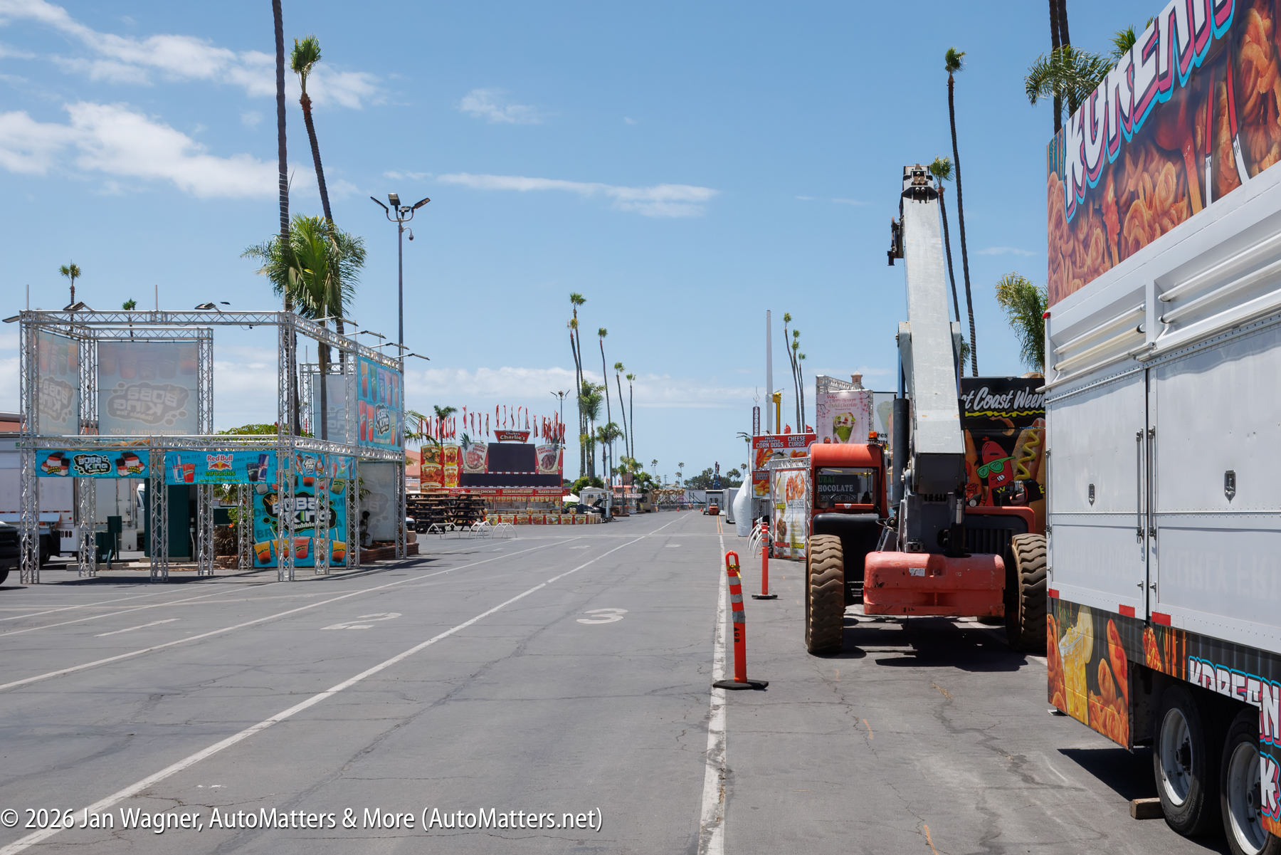 A fairground street with food trucks and equipment being set up, palm trees lining the area, and clear blue sky overhead.