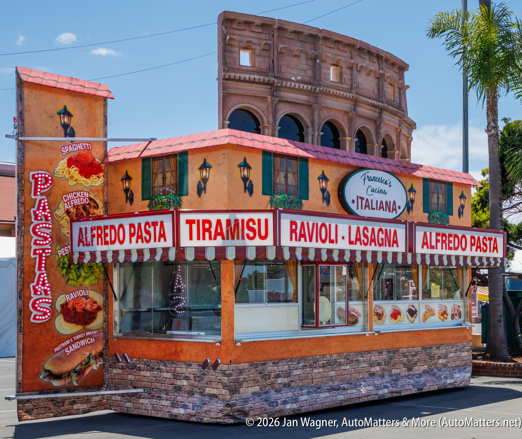 A food stand with an Italian décor advertises Alfredo pasta, tiramisu, ravioli, and lasagna, with large signs and images of menu items. A replica of the Colosseum is in the background.