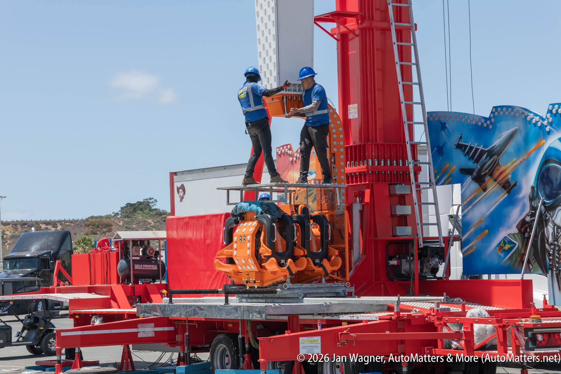 Two workers in blue helmets install orange seats on a red amusement park ride structure outdoors, with a mural and trucks in the background.