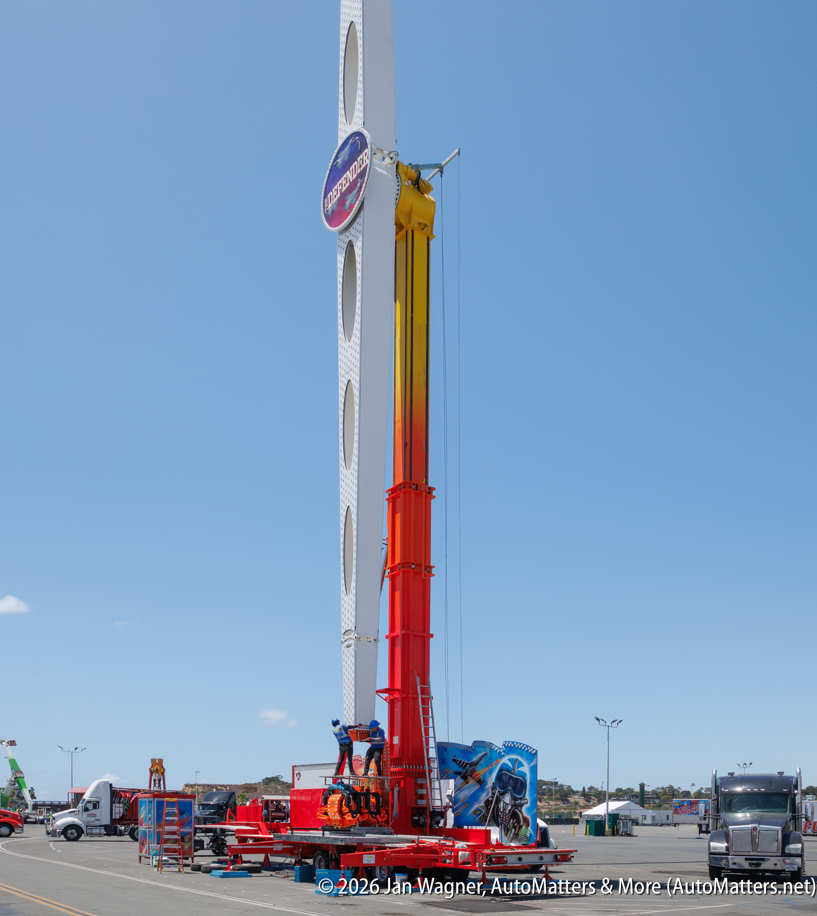 A tall amusement park ride with a rotating arm and circular logo stands upright in a parking lot under a clear blue sky.