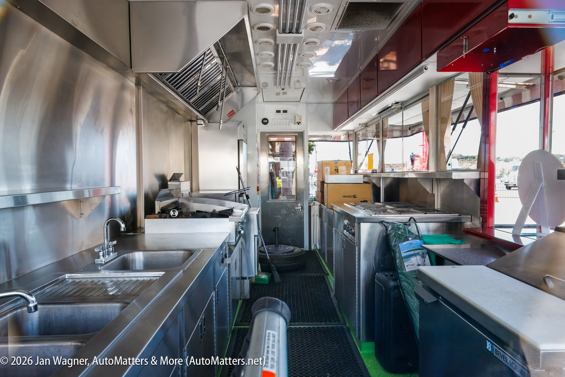 Interior of a clean, modern food truck kitchen with stainless steel counters, sinks, and cooking equipment, with supplies and boxes visible throughout.