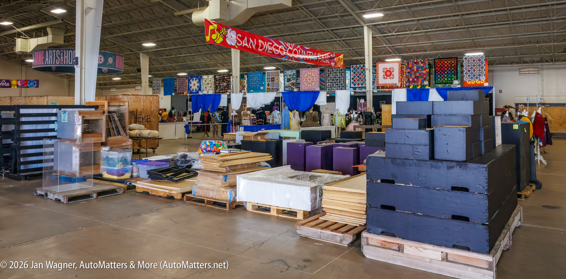 Stacks of boxes, pallets, and furniture are arranged in a large indoor hall with quilts displayed on the back wall and a "San Diego" banner above.