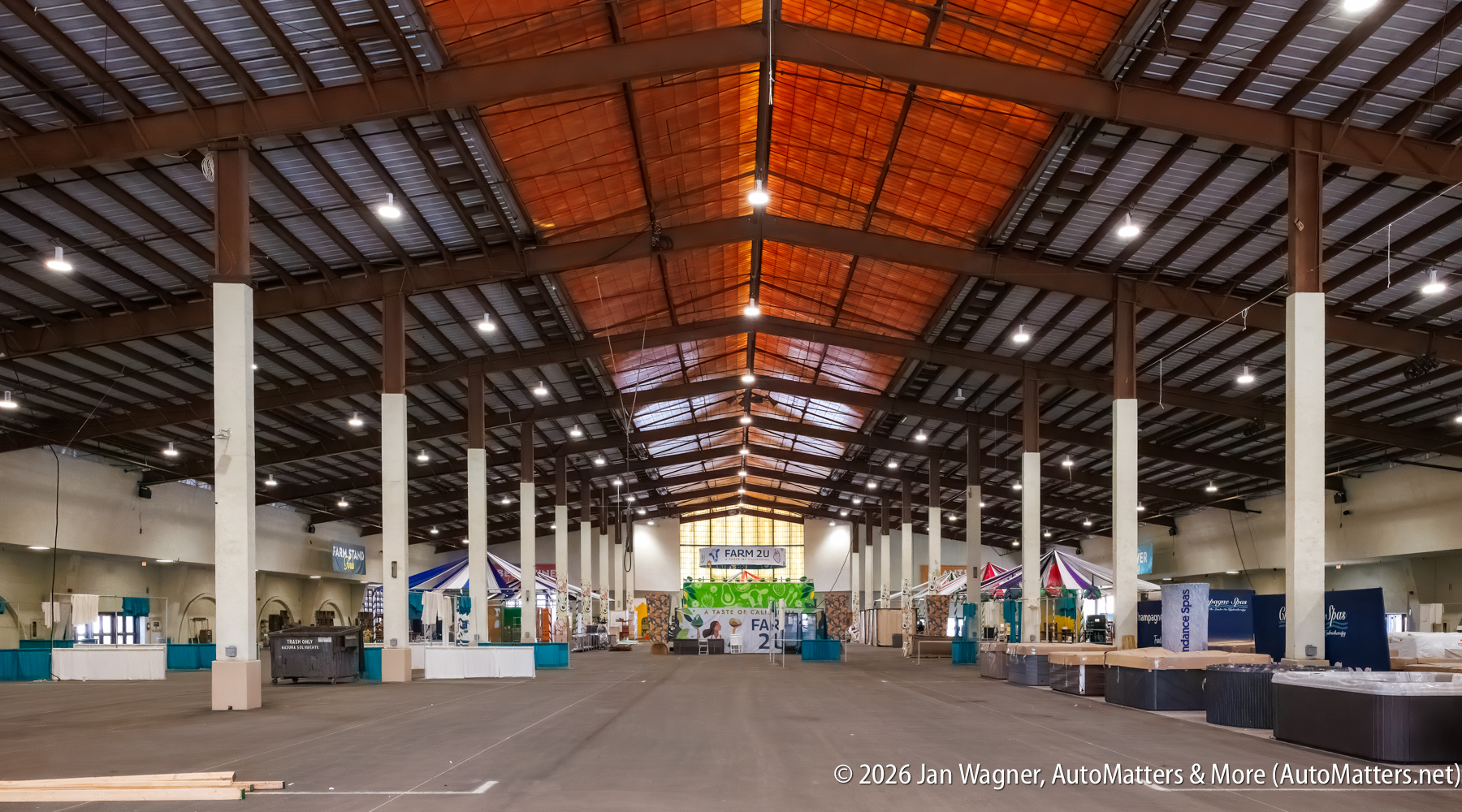 Large indoor exhibition hall with high ceilings, pillars, and various colorful booths being set up along the sides; open space in the center.