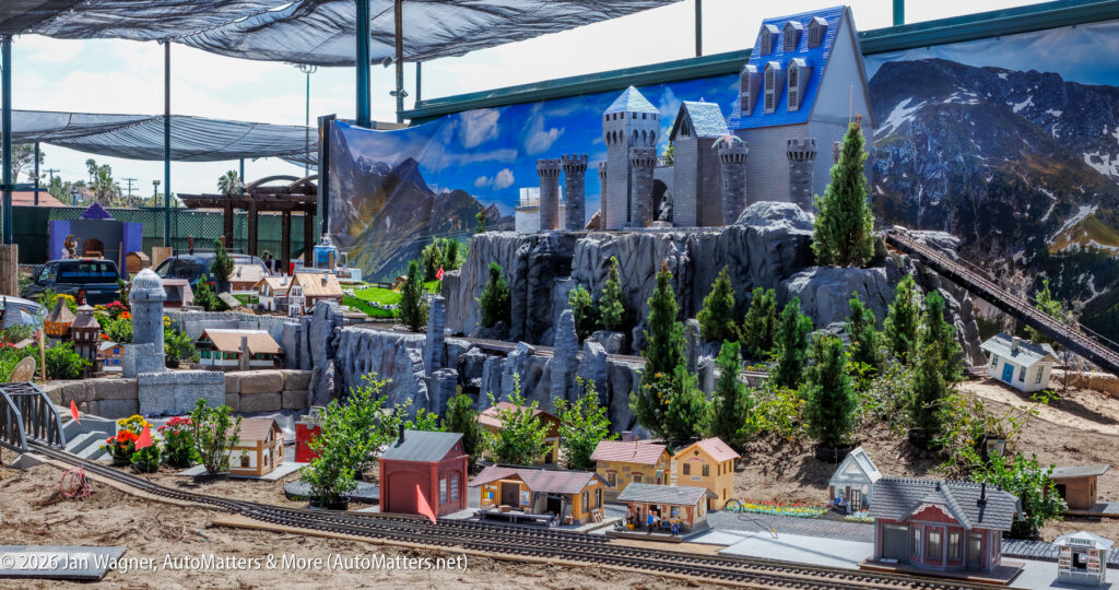 Model train set with miniature houses, a castle on a rocky hill, small trees, and painted mountain backdrop under netted shade.