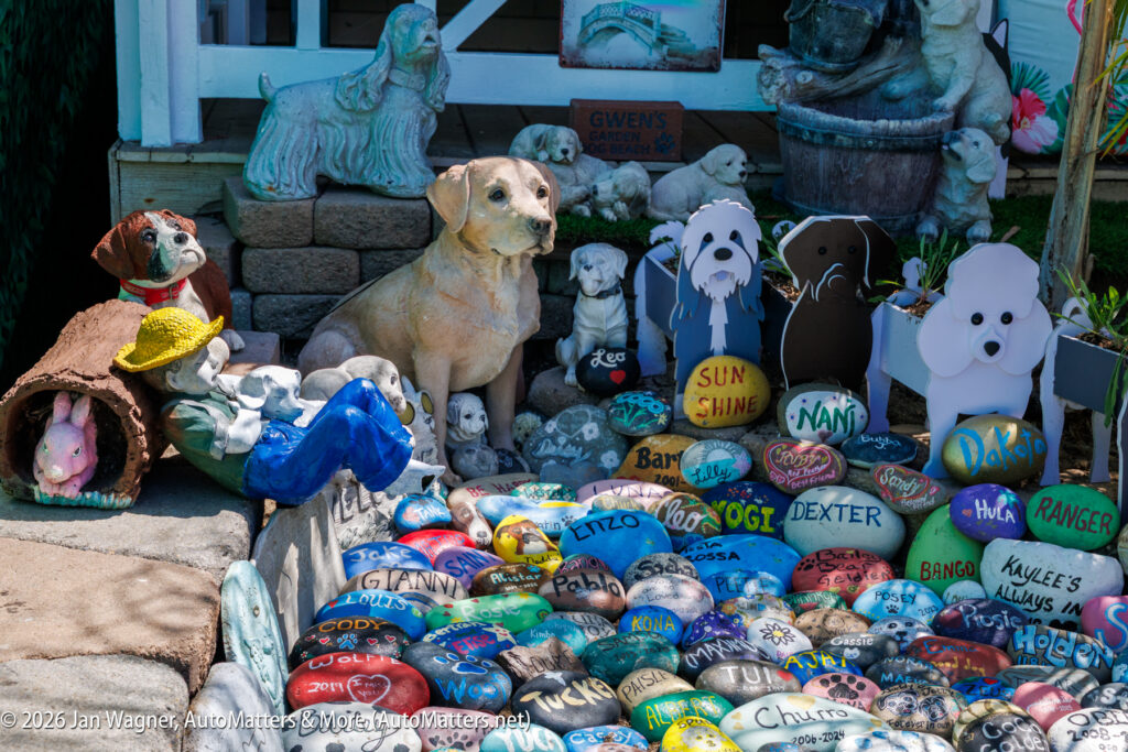 A collection of painted rocks with pet names, surrounded by dog statues and cutouts, displayed outdoors near a porch.