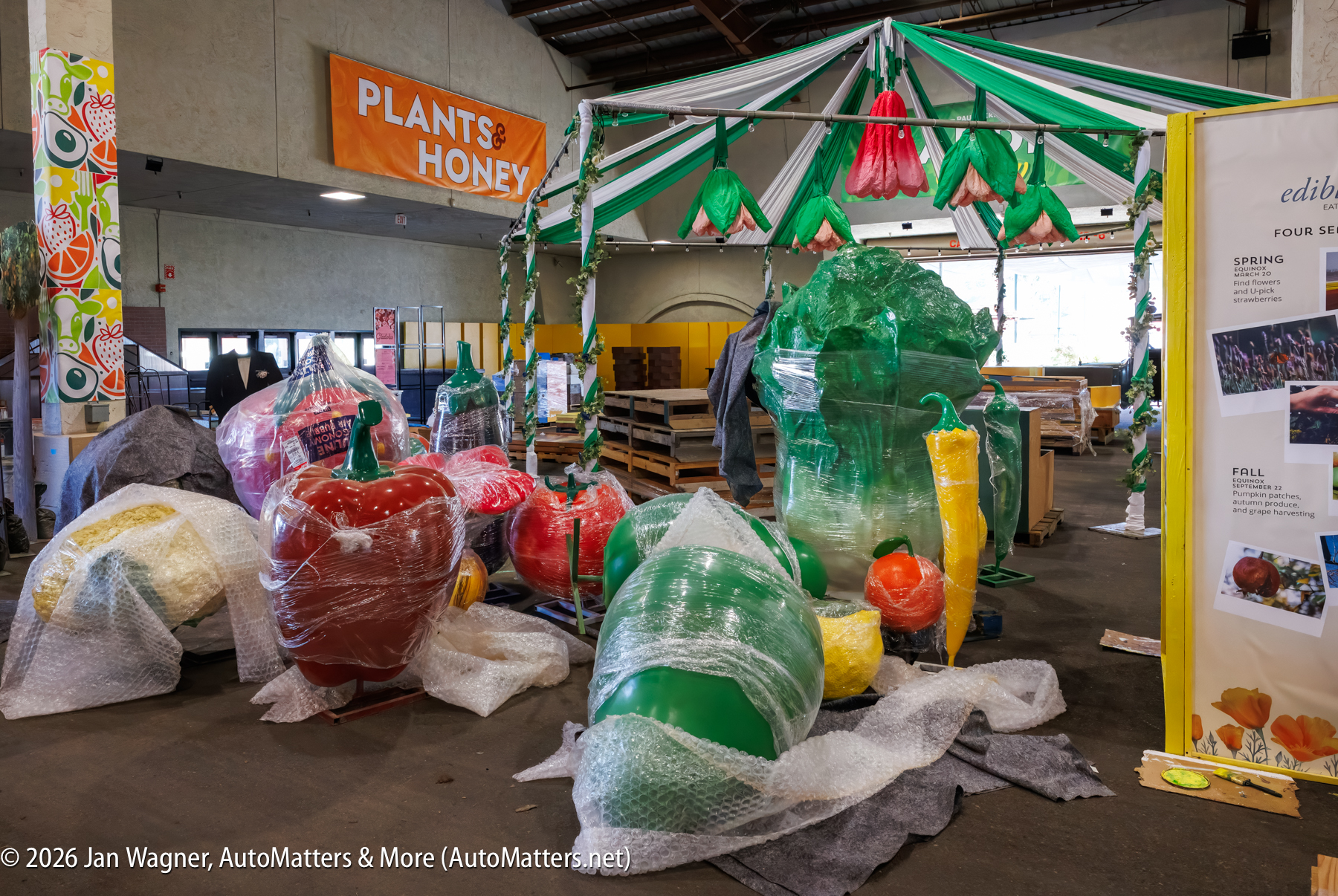 Large plastic models of fruits and vegetables wrapped in bubble wrap are displayed inside a warehouse decorated with a “Plants & Honey” sign and colorful banners.