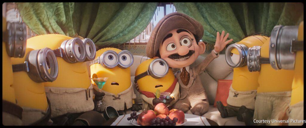 Animated character in a hat and mustache sits on a couch, waving, surrounded by five Minions, with fruit and grapes on the table in front of them.