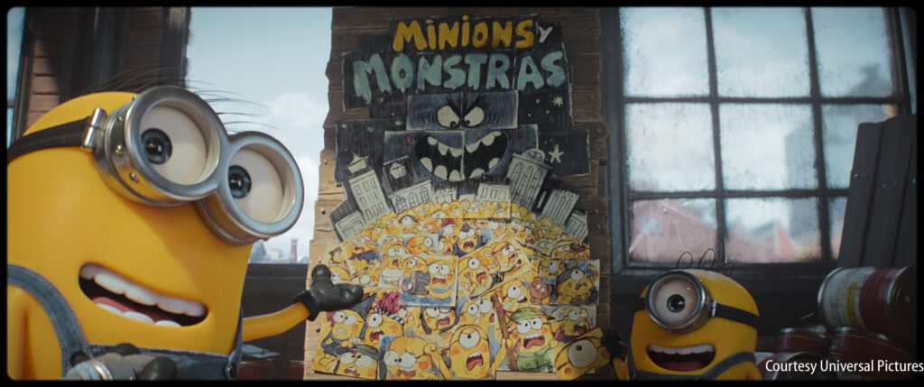 Two Minions stand beside a colorful poster titled "Minions y Monstras," showing many Minions in front of a large cartoonish monster.