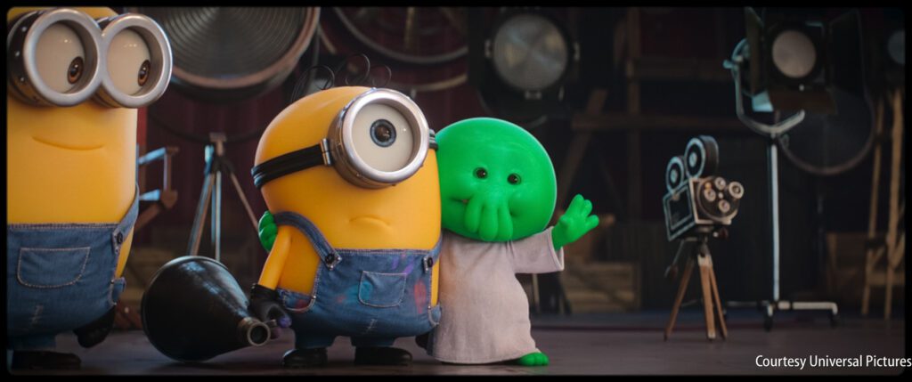 Two Minions in overalls stand beside a small green creature waving, with film equipment visible in the background.