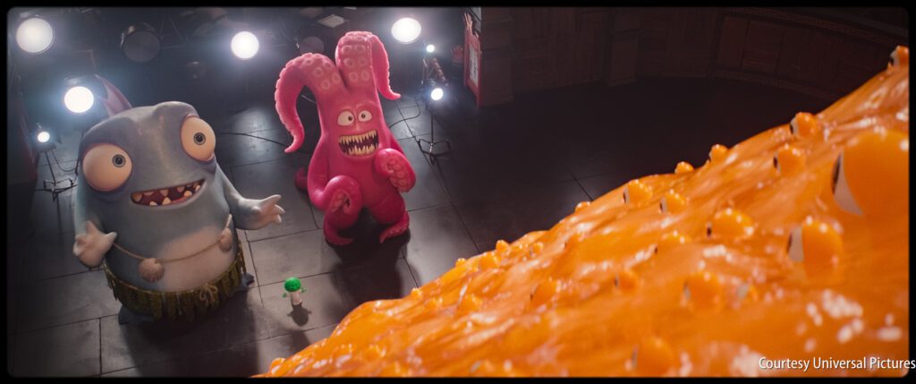 Two cartoonish monsters, one blue and one pink, stand on a stage looking up at a large orange blob under bright lights.