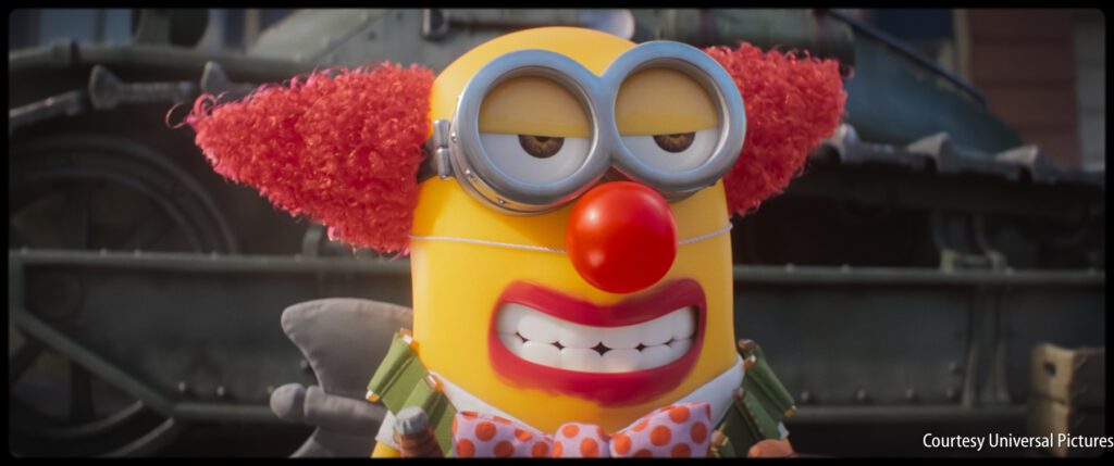 A Minion dressed as a clown with red hair, big glasses, and a red nose stands in front of a tank, wearing a polka-dot bow tie and grimacing.