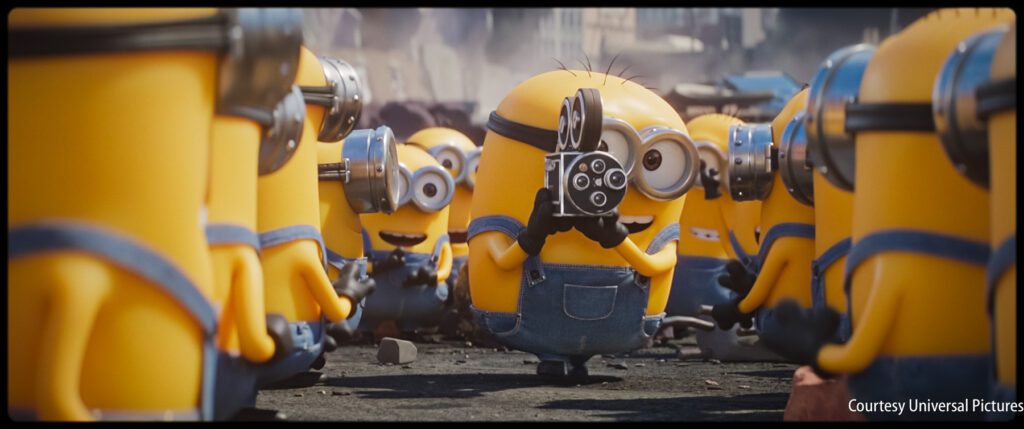 Several yellow, goggle-wearing Minions gather outdoors; one Minion in the center is filming with an old-fashioned movie camera.