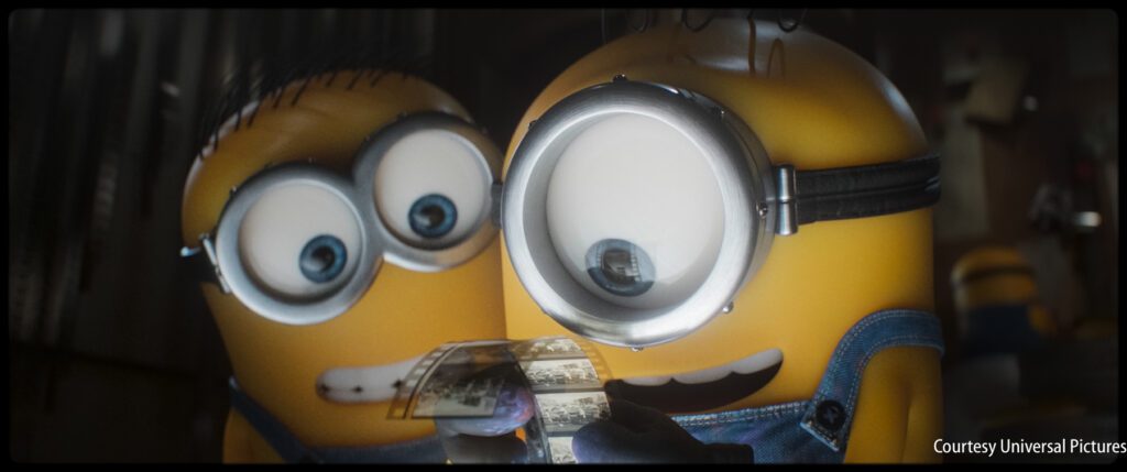Two Minions examine a strip of photographic film closely; one holds the film while the other looks over his shoulder.
